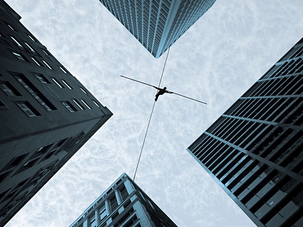 view looking up at a person walking a tightrope between tall buildings 